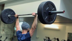 a man wearing a beanie and overhead pressing a loaded barbell in a gym setting