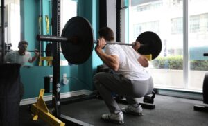 a man wearing Olympic lifting shoes performing a high-bar squat with a loaded barbell next to a rack inside a gym